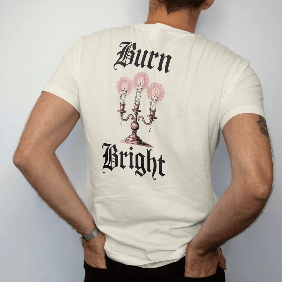 Man wearing a Ivory tee, seen from the back: back print: ornate candelabra with three dripping candles and a moth circling the flames, Victorian engraving with stippled glow, "BURN" above in ornate gothic blackletter, "BRIGHT" below in thin…
