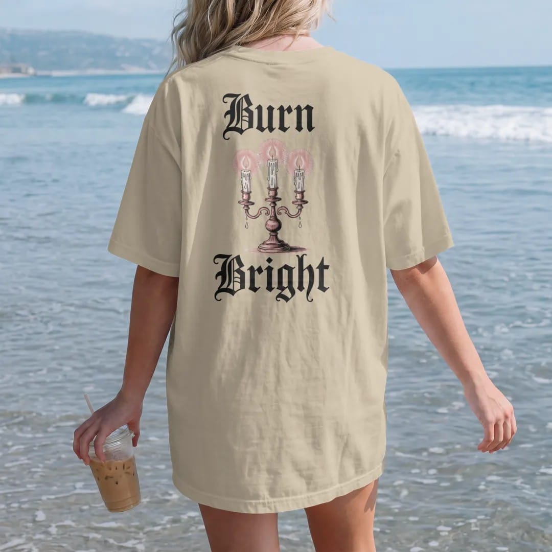 Woman on the beach in a Sandstone tee, back view: back print: ornate candelabra with three dripping candles and a moth circling the flames, Victorian engraving with stippled glow, "BURN" above in ornate gothic blackletter, "BRIGHT" below in thin…