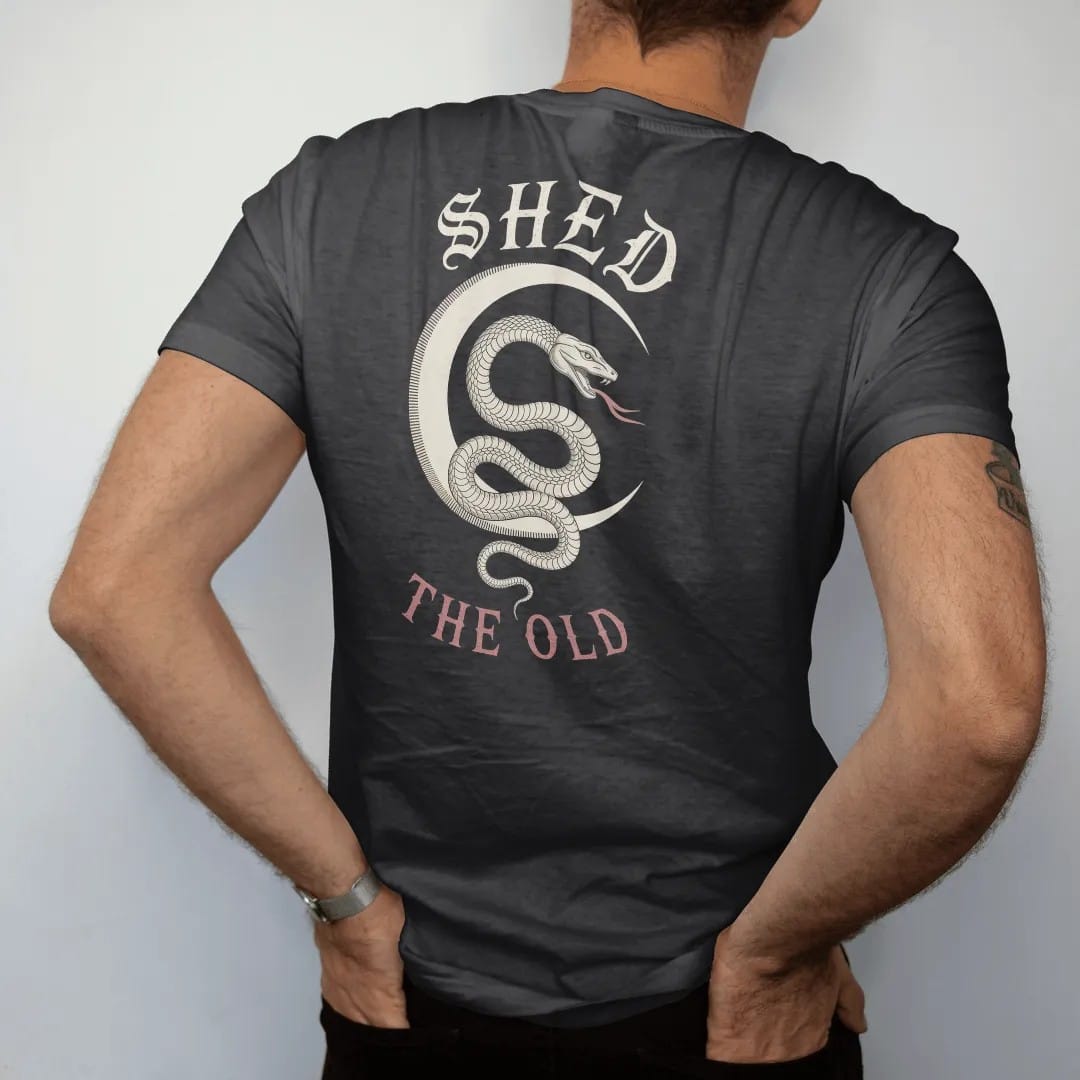 Man wearing a Black tee, seen from the back: back print: serpent coiled around a crescent moon with a shed skin trailing below, fine engraving with crosshatching, "SHED" above in ornate gothic blackletter, "THE OLD" below in small tracked engraved…