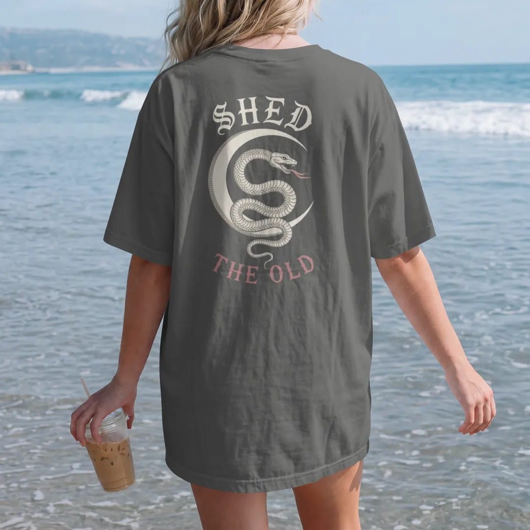 Woman on the beach in a Pepper tee, back view: back print: serpent coiled around a crescent moon with a shed skin trailing below, fine engraving with crosshatching, "SHED" above in ornate gothic blackletter, "THE OLD" below in small tracked…