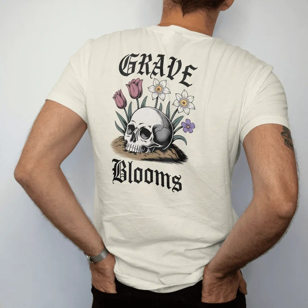 Man wearing a Ivory tee, seen from the back: back print: skull half buried in soil with spring tulips, daffodils and violets sprouting through its eye sockets, antique engraving style, "GRAVE" above in ornate gothic blackletter, "BLOOMS" below in…