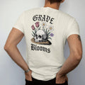 Man wearing a Ivory tee, seen from the back: back print: skull half buried in soil with spring tulips, daffodils and violets sprouting through its eye sockets, antique engraving style, "GRAVE" above in ornate gothic blackletter, "BLOOMS" below in…