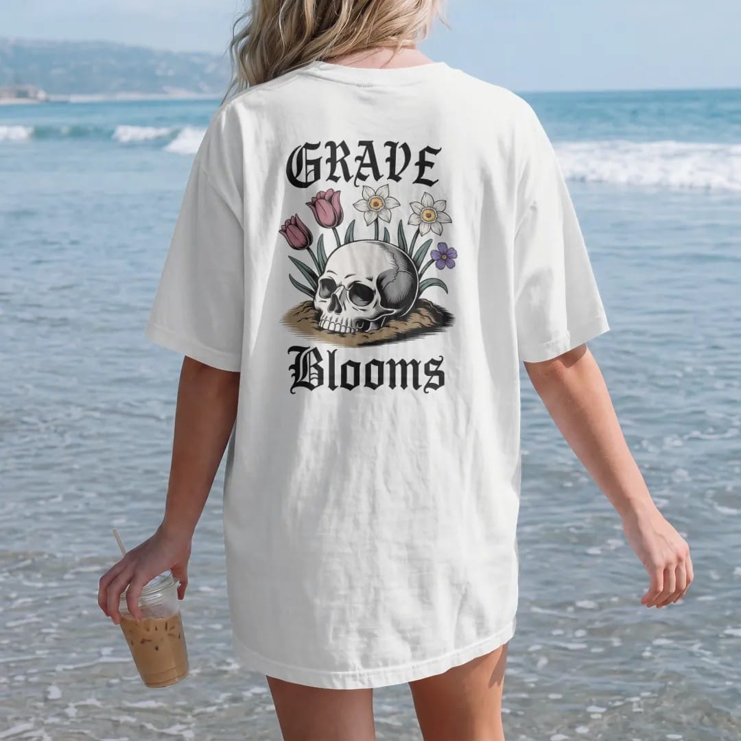 Woman on the beach in a White tee, back view: back print: skull half buried in soil with spring tulips, daffodils and violets sprouting through its eye sockets, antique engraving style, "GRAVE" above in ornate gothic blackletter, "BLOOMS" below in…