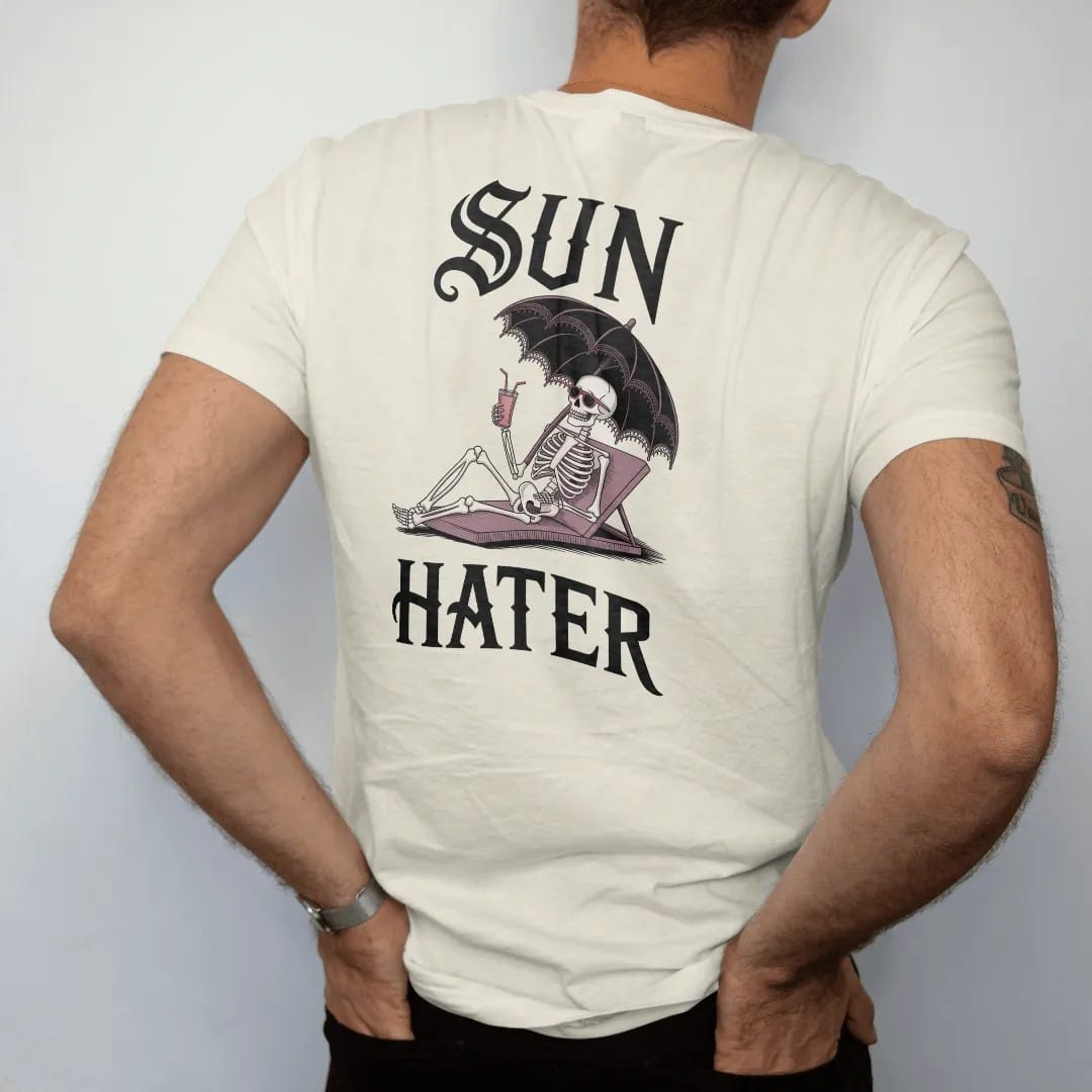 Man wearing a Ivory tee, seen from the back: back print: skeleton lounging under a black lace parasol with sunglasses and a cold drink, Victorian engraving with a wry twist, "SUN" above in ornate gothic blackletter, "HATER" below in elegant…