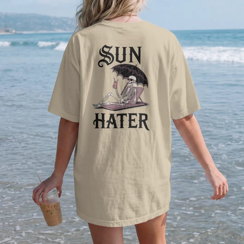 Woman on the beach in a Sandstone tee, back view: back print: skeleton lounging under a black lace parasol with sunglasses and a cold drink, Victorian engraving with a wry twist, "SUN" above in ornate gothic blackletter, "HATER" below in elegant…