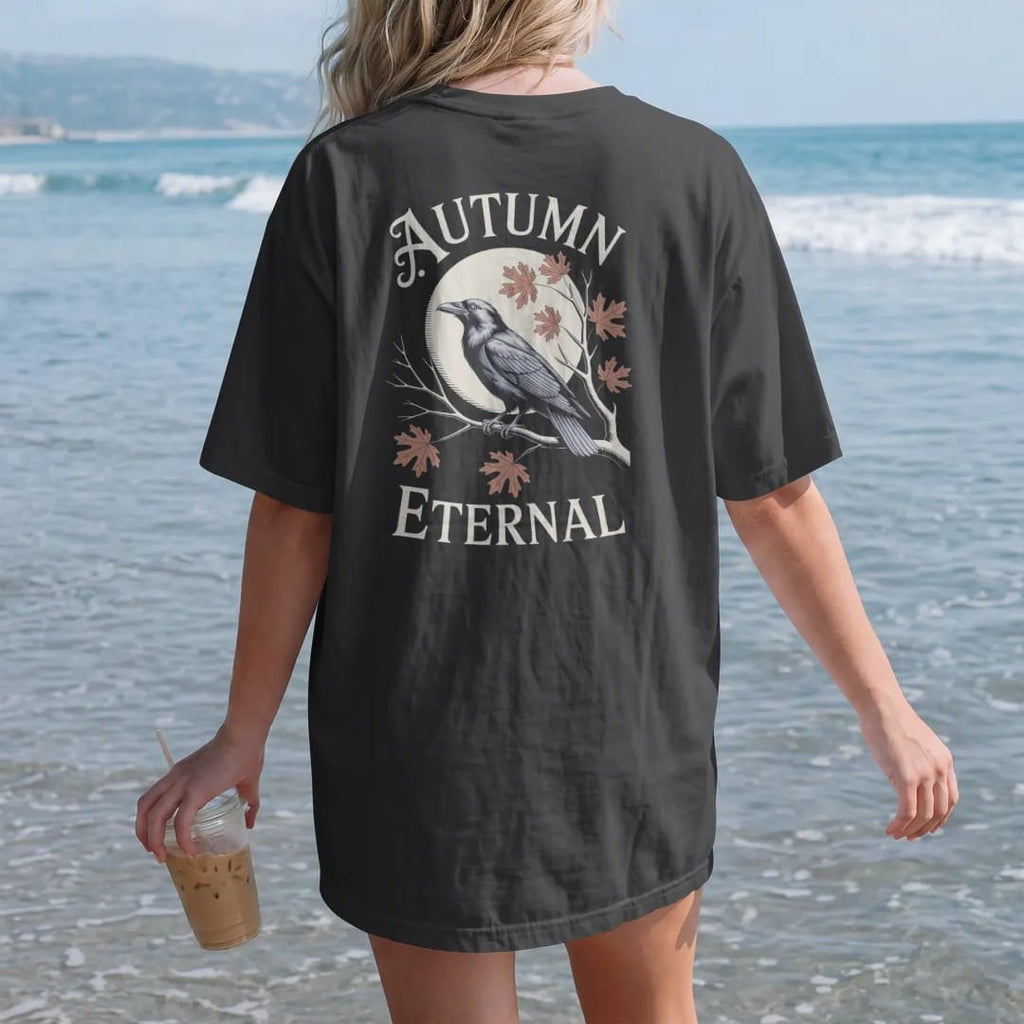 Woman on the beach in a Black tee, back view: back print: raven on a bare branch with oak and maple leaves drifting down past a harvest moon, fine engraving style, "AUTUMN" above in ornate gothic blackletter, "ETERNAL" below in small tracked…