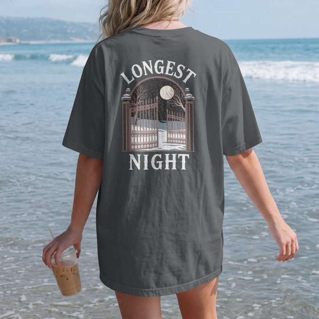 Woman on the beach in a Graphite tee, back view: back print: wrought-iron cemetery gate under falling snow with a full moon and bare trees, detailed Victorian engraving, "LONGEST" arched above in ornate gothic blackletter, "NIGHT" below in elegant…