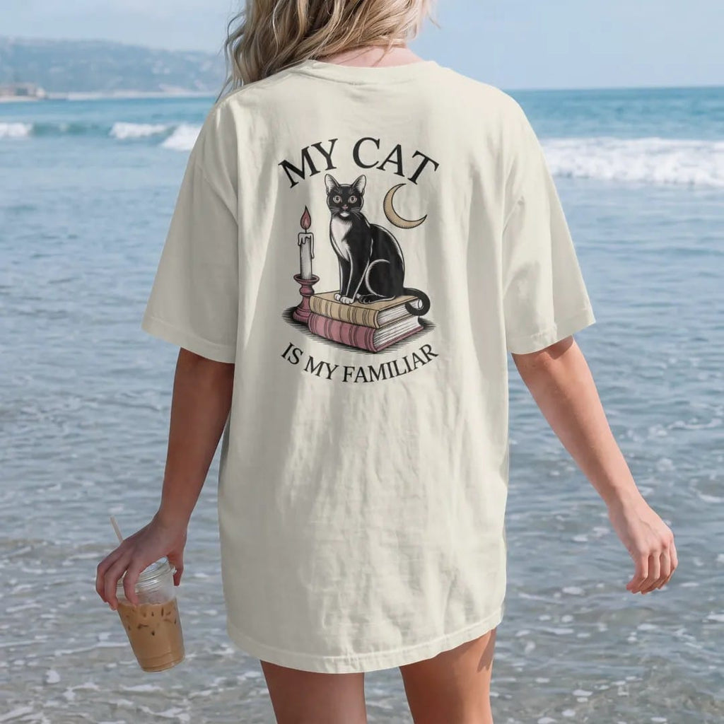 Woman on the beach in a Ivory tee, back view: back print: black cat sitting on a stack of old books with a crescent moon and candle, fine Victorian engraving style, "MY CAT" above in elegant high-contrast serif caps, "IS MY FAMILIAR" below in thin…