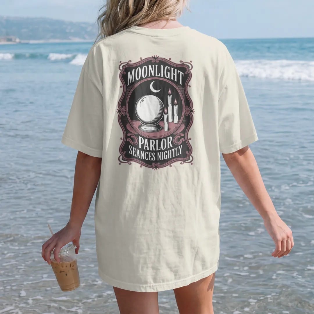 Woman on the beach in a Ivory tee, back view: back print: victorian theatre handbill with a crystal ball, candles and a crescent moon, ornate playbill border, detailed vintage engraving with fine crosshatching, "MOONLIGHT" in elegant serif caps,…
