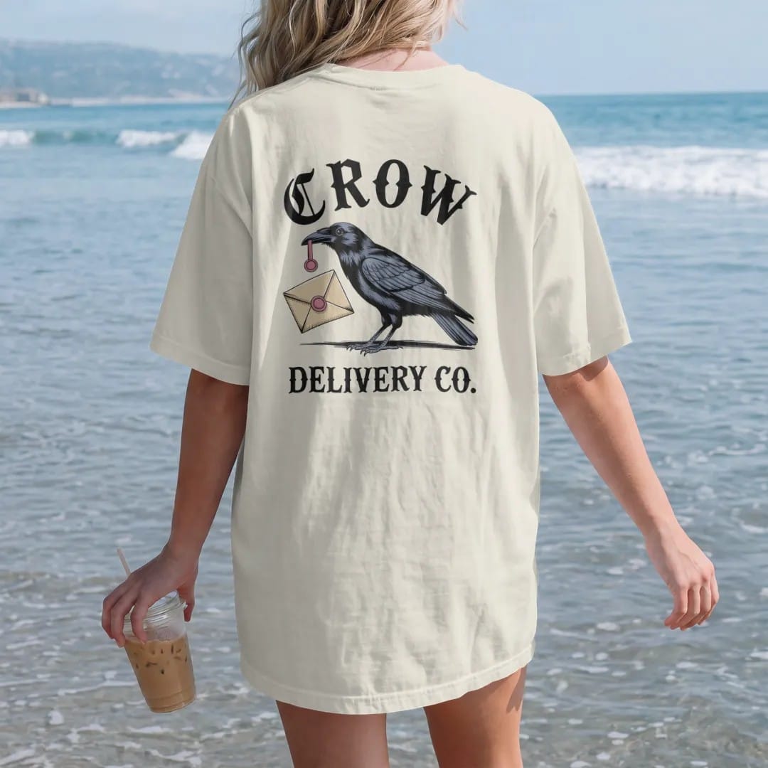 Woman on the beach in a Ivory tee, back view: back print: crow carrying a sealed envelope with a wax seal in its beak, vintage postal advertisement style engraving, "CROW" arched above in ornate gothic blackletter, "DELIVERY CO." below in small…