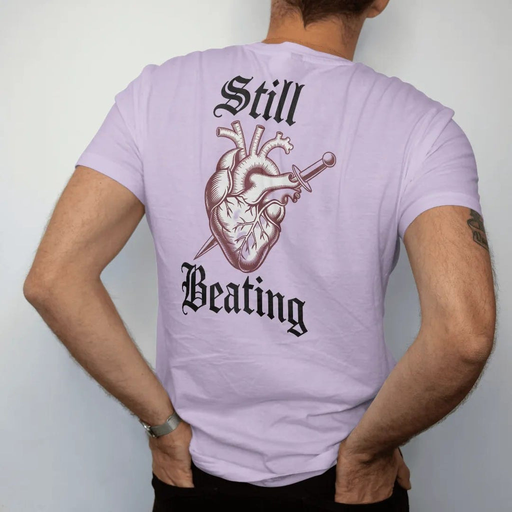 Man wearing a Orchid tee, seen from the back: back print: anatomical heart wrapped in thorny roses with a small dagger through it, antique medical engraving style, "STILL" above in ornate gothic blackletter, "BEATING" below in thin copperplate…