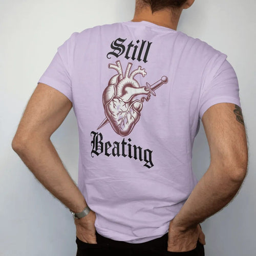 Man wearing a Orchid tee, seen from the back: back print: anatomical heart wrapped in thorny roses with a small dagger through it, antique medical engraving style, "STILL" above in ornate gothic blackletter, "BEATING" below in thin copperplate…