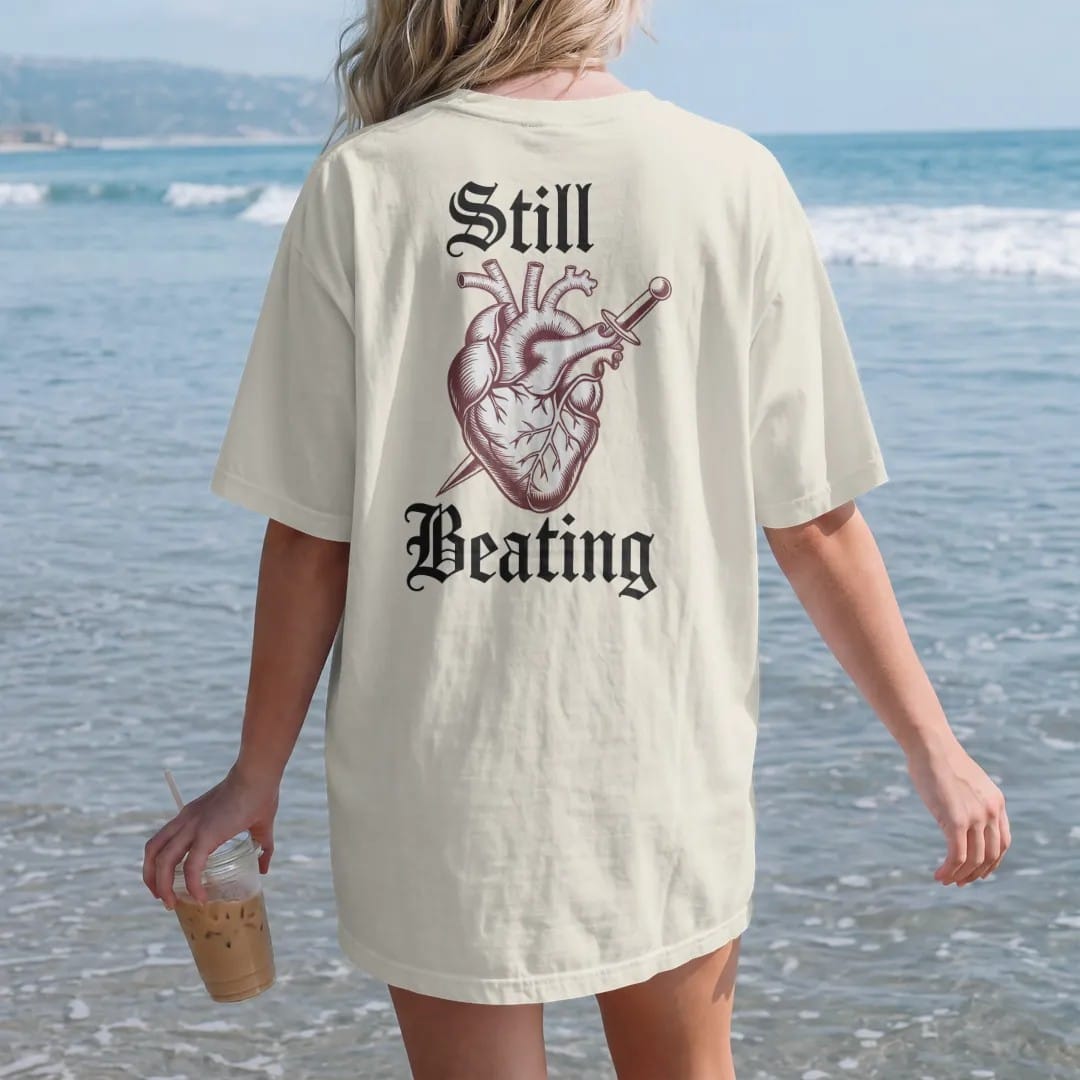Woman on the beach in a Ivory tee, back view: back print: anatomical heart wrapped in thorny roses with a small dagger through it, antique medical engraving style, "STILL" above in ornate gothic blackletter, "BEATING" below in thin copperplate…