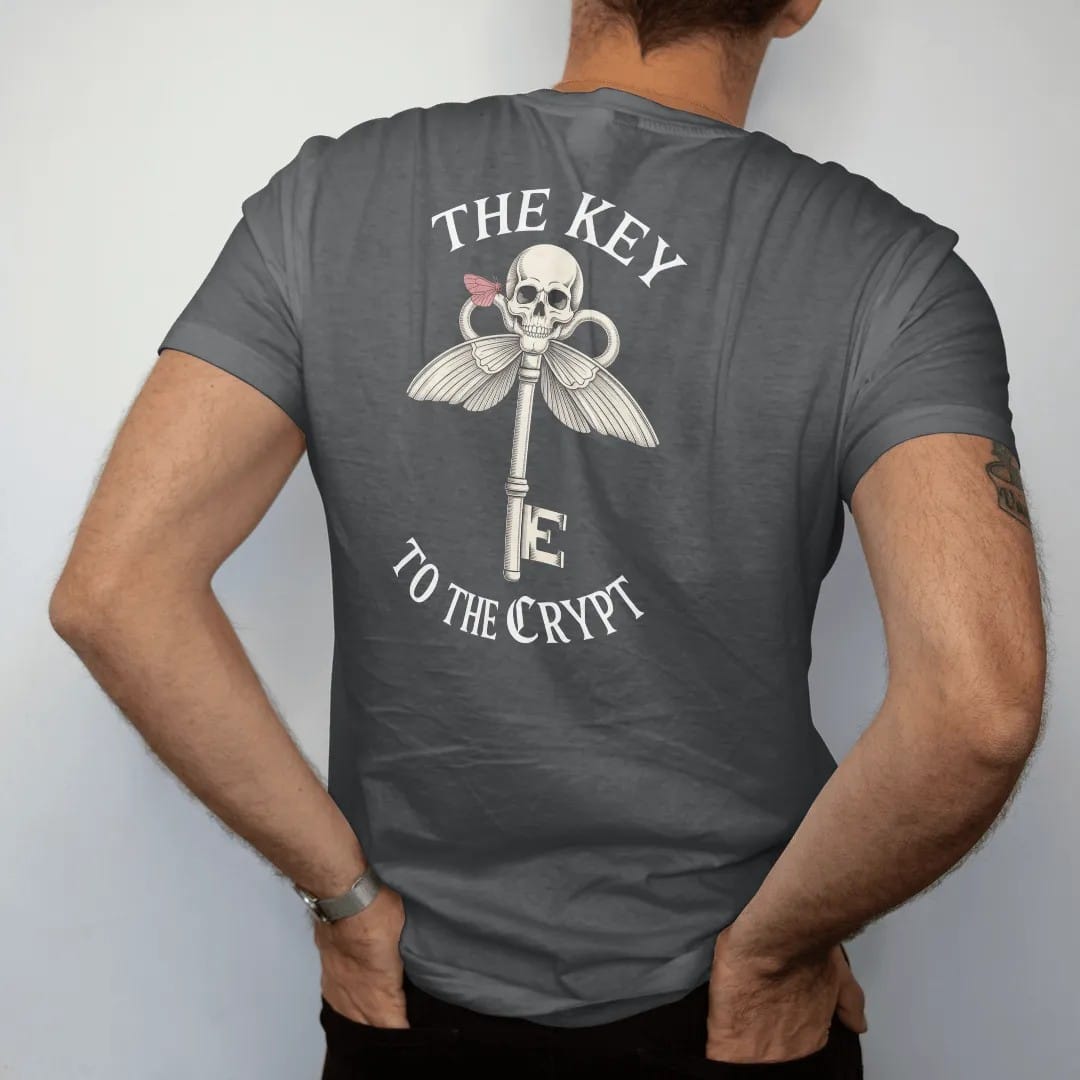 Man wearing a Graphite tee, seen from the back: back print: ornate skeleton key with a skull bow and a moth perched on it, Victorian engraving with crosshatching, "THE KEY" above in elegant high-contrast serif caps, "TO THE CRYPT" below in ornate…