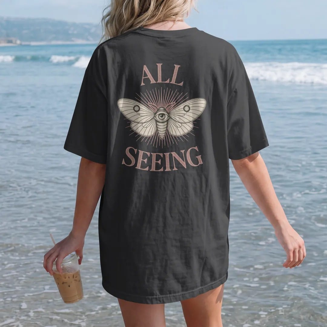 Woman on the beach in a Black tee, back view: back print: single open eye with moth wings spreading from it and rays behind, mystical engraving style, "ALL" above in elegant high-contrast serif caps, "SEEING" below in small tracked engraved caps,…