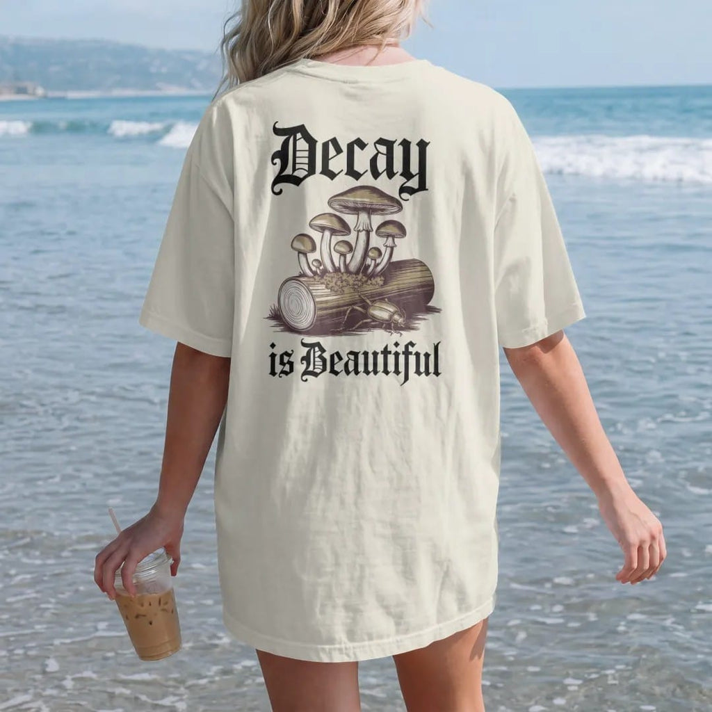 Woman on the beach in a Ivory tee, back view: back print: cluster of wild mushrooms and moss growing from a fallen log with a beetle, antique botanical engraving, "DECAY" above in ornate gothic blackletter, "IS BEAUTIFUL" below in thin copperplate…