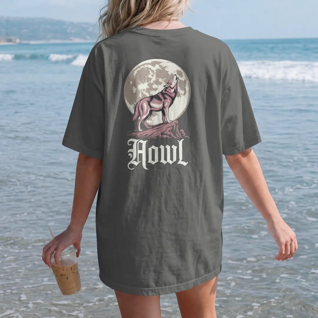 Woman on the beach in a Pepper tee, back view: back print: lone wolf howling on a rocky ridge in front of a huge full moon, detailed Victorian engraving with stippling, "HOWL" below in ornate gothic blackletter, printed in bone cream, antique gold…