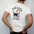 Man wearing a White tee, seen from the back: back print: black cat walking under a ladder past a broken mirror, wry Victorian engraving style, "BAD LUCK" arched above in ornate gothic blackletter, "CLUB" below in elegant high-contrast serif caps,…
