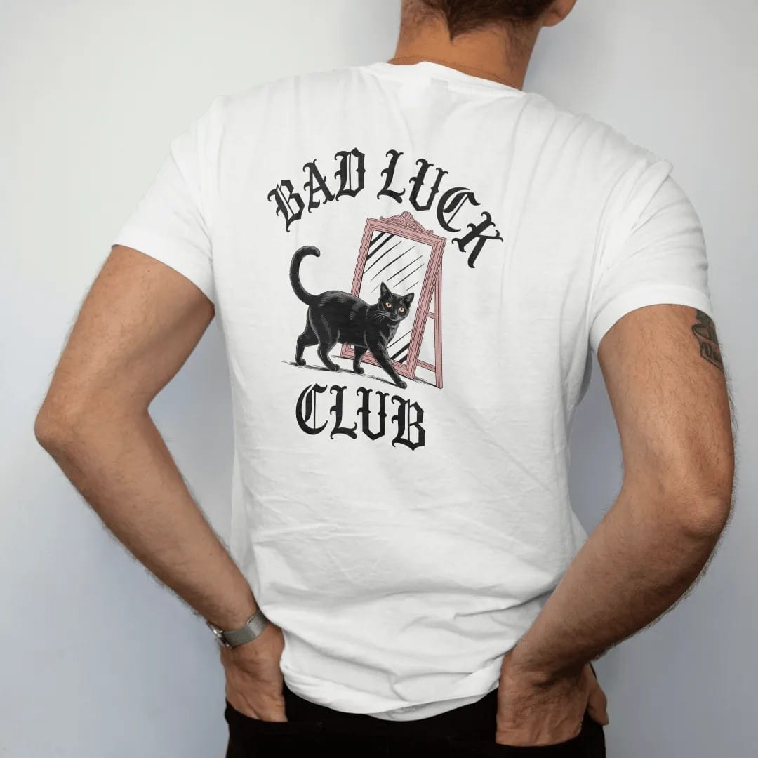 Man wearing a White tee, seen from the back: back print: black cat walking under a ladder past a broken mirror, wry Victorian engraving style, "BAD LUCK" arched above in ornate gothic blackletter, "CLUB" below in elegant high-contrast serif caps,…