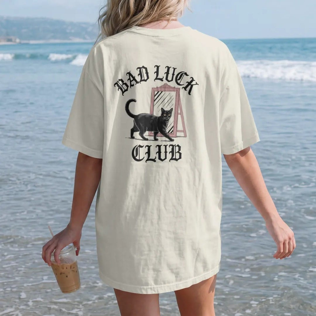 Woman on the beach in a Ivory tee, back view: back print: black cat walking under a ladder past a broken mirror, wry Victorian engraving style, "BAD LUCK" arched above in ornate gothic blackletter, "CLUB" below in elegant high-contrast serif caps,…