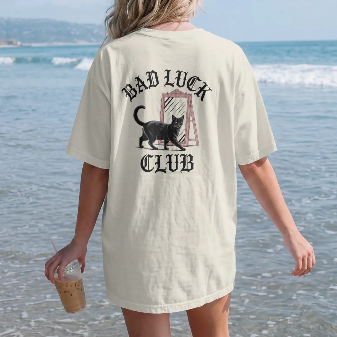 Woman on the beach in a Ivory tee, back view: back print: black cat walking under a ladder past a broken mirror, wry Victorian engraving style, "BAD LUCK" arched above in ornate gothic blackletter, "CLUB" below in elegant high-contrast serif caps,…