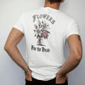Man wearing a White tee, seen from the back: back print: bouquet of lilies and roses tied with ribbon, stems wrapped around small bones, Victorian mourning-card engraving style, "FLOWERS" above in thin copperplate script, "FOR THE DEAD" below in…