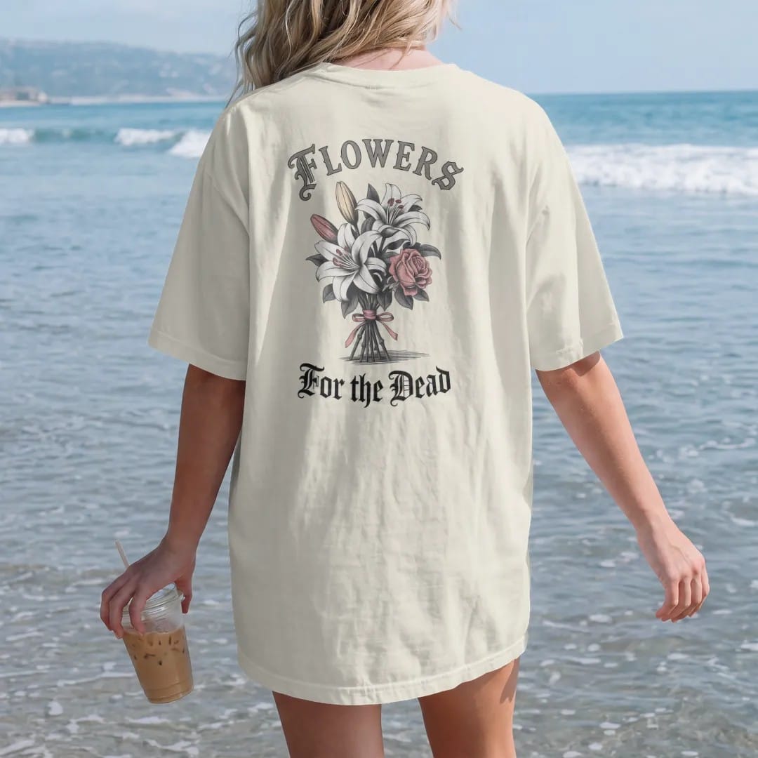 Woman on the beach in a Ivory tee, back view: back print: bouquet of lilies and roses tied with ribbon, stems wrapped around small bones, Victorian mourning-card engraving style, "FLOWERS" above in thin copperplate script, "FOR THE DEAD" below in…