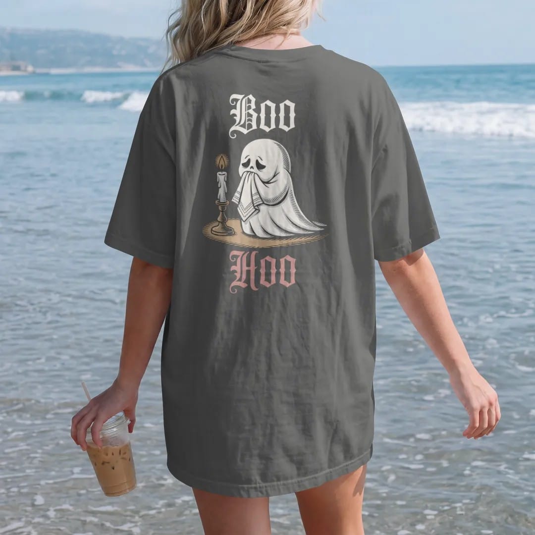 Woman on the beach in a Pepper tee, back view: back print: little sheet ghost crying into a lace handkerchief by candlelight, playful Victorian engraving style, "BOO" above in ornate gothic blackletter, "HOO" below in thin copperplate script,…