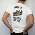 Man wearing a White tee, seen from the back: back print: skeleton lying in an open coffin holding a steaming cup of coffee, wry Victorian engraving style, "NOT A" above in elegant high-contrast serif caps, "MORNING PERSON" below in ornate gothic…