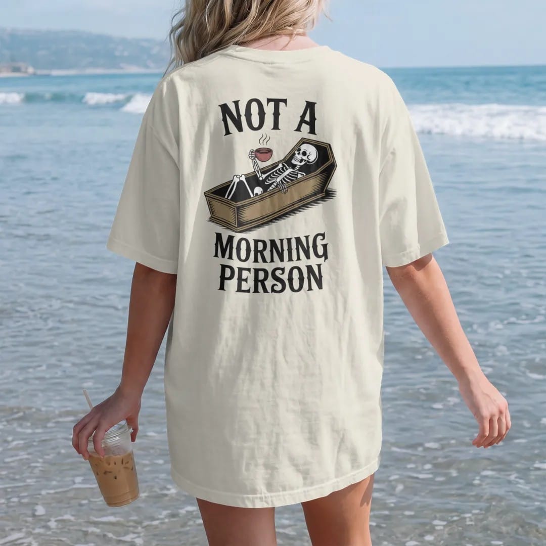Woman on the beach in a Ivory tee, back view: back print: skeleton lying in an open coffin holding a steaming cup of coffee, wry Victorian engraving style, "NOT A" above in elegant high-contrast serif caps, "MORNING PERSON" below in ornate gothic…