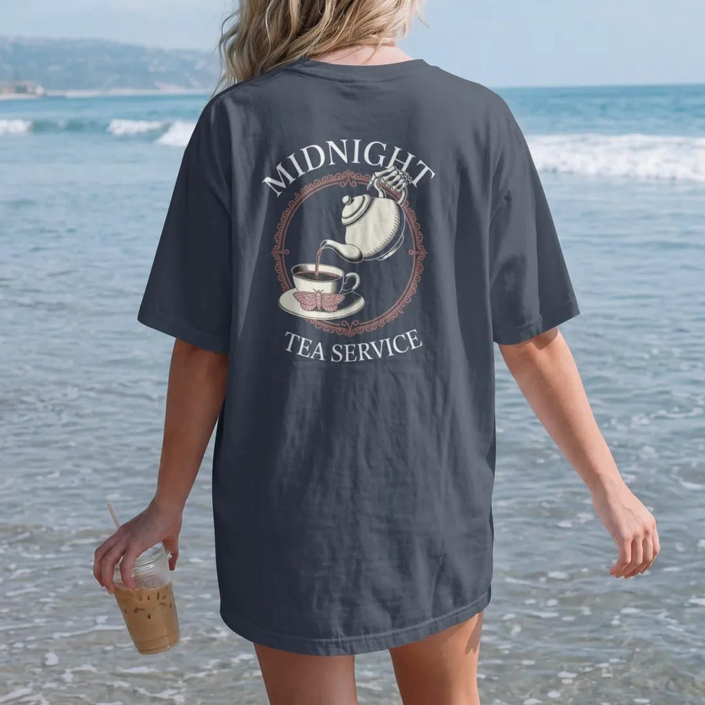 Woman on the beach in a Midnight tee, back view: back print: skeleton hand pouring tea from an ornate teapot into a cup with a moth on the saucer, Victorian engraving, "MIDNIGHT" above in elegant high-contrast serif caps, "TEA SERVICE" below in…