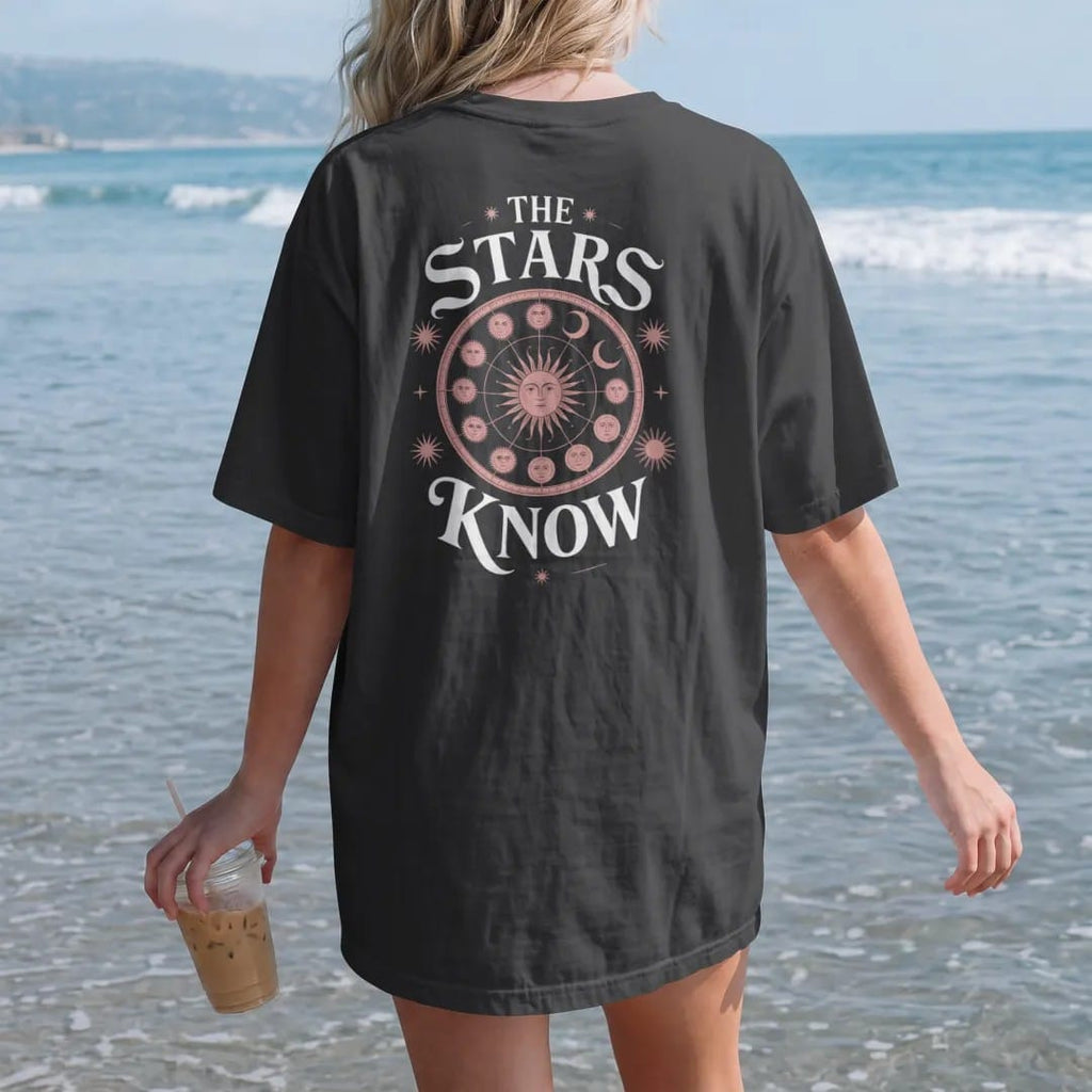 Woman on the beach in a Black tee, back view: back print: antique zodiac wheel with constellations, sun and moon faces in a celestial engraving style, "THE" small above, "STARS" in ornate gothic blackletter, "KNOW" below in elegant high-contrast…