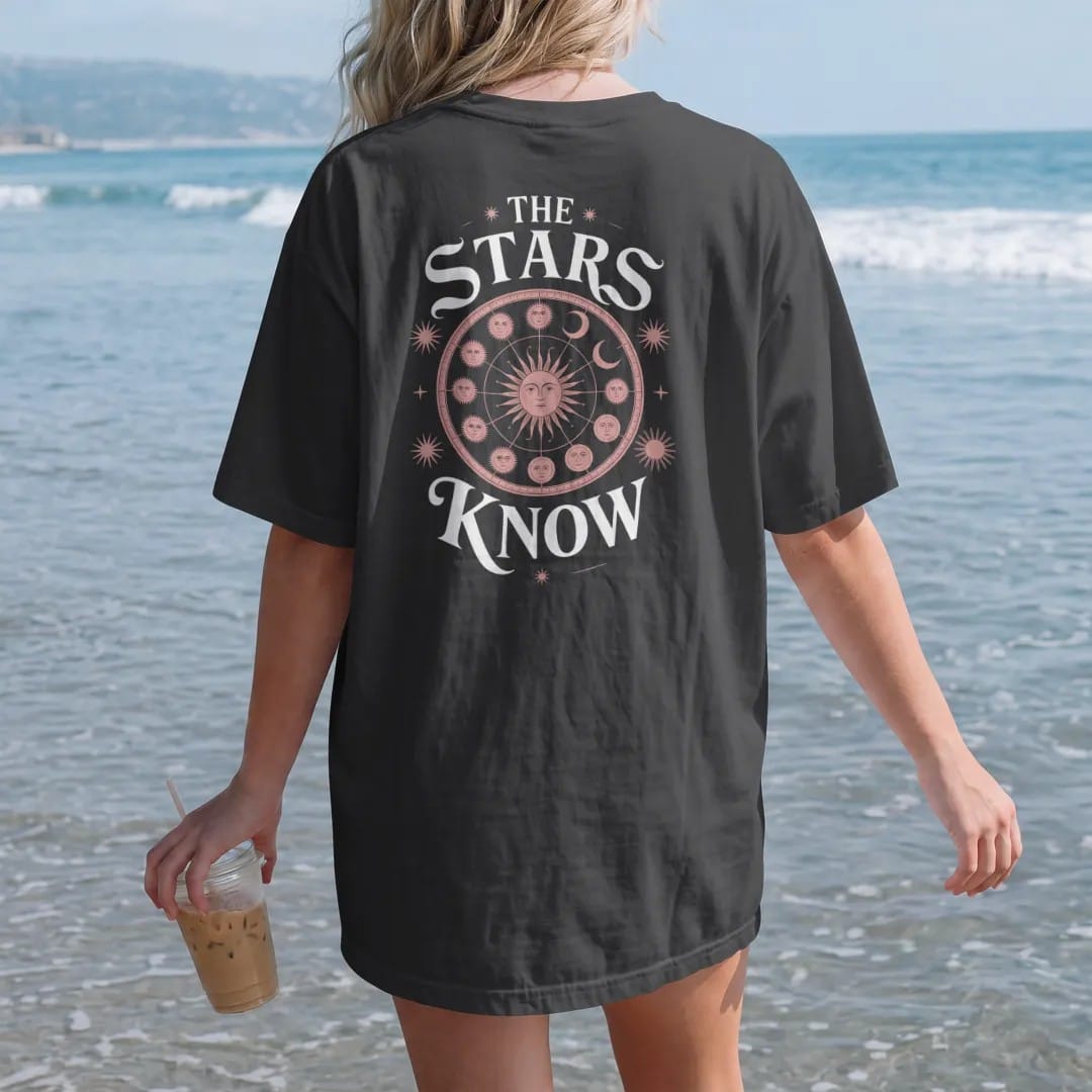 Woman on the beach in a Black tee, back view: back print: antique zodiac wheel with constellations, sun and moon faces in a celestial engraving style, "THE" small above, "STARS" in ornate gothic blackletter, "KNOW" below in elegant high-contrast…