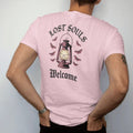 Man wearing a Blossom tee, seen from the back: back print: old iron lantern glowing on a hook with moths circling the light, Victorian engraving with stippled glow, "LOST SOULS" arched above in ornate gothic blackletter, "WELCOME" below in thin…
