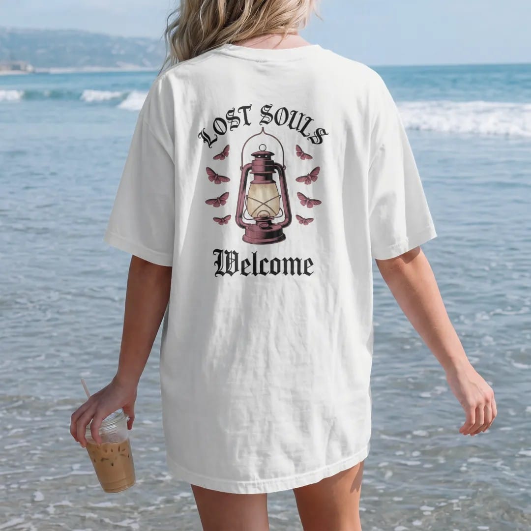 Woman on the beach in a White tee, back view: back print: old iron lantern glowing on a hook with moths circling the light, Victorian engraving with stippled glow, "LOST SOULS" arched above in ornate gothic blackletter, "WELCOME" below in thin…