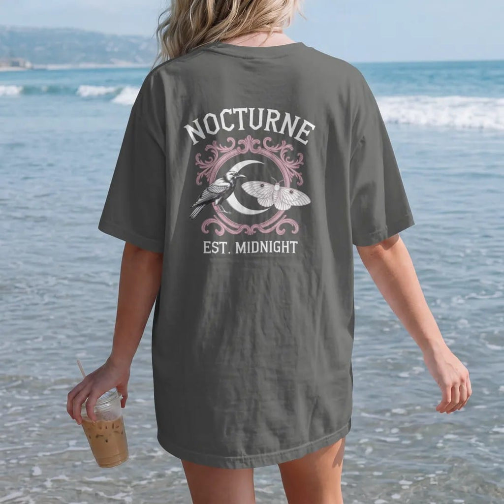 Woman on the beach in a Pepper tee, back view: back print: heraldic crest with a raven and a moth flanking a crescent moon shield, ornate Victorian engraving, "NOCTURNE" arched above in ornate gothic blackletter, "EST. MIDNIGHT" below in small…
