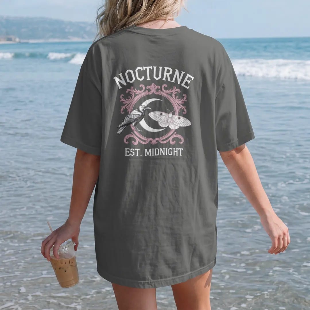 Woman on the beach in a Pepper tee, back view: back print: heraldic crest with a raven and a moth flanking a crescent moon shield, ornate Victorian engraving, "NOCTURNE" arched above in ornate gothic blackletter, "EST. MIDNIGHT" below in small…