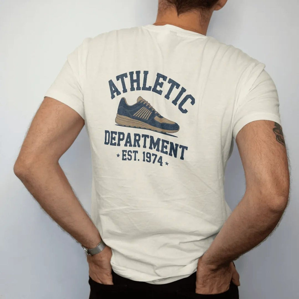 Man wearing a Ivory tee, seen from the back: a vintage running shoe with speed lines, with "ATHLETIC" and "DEPARTMENT" and "EST. 1974" in chunky faded collegiate block letters, printed in faded varsity navy, washed red, mustard gold and pine green…