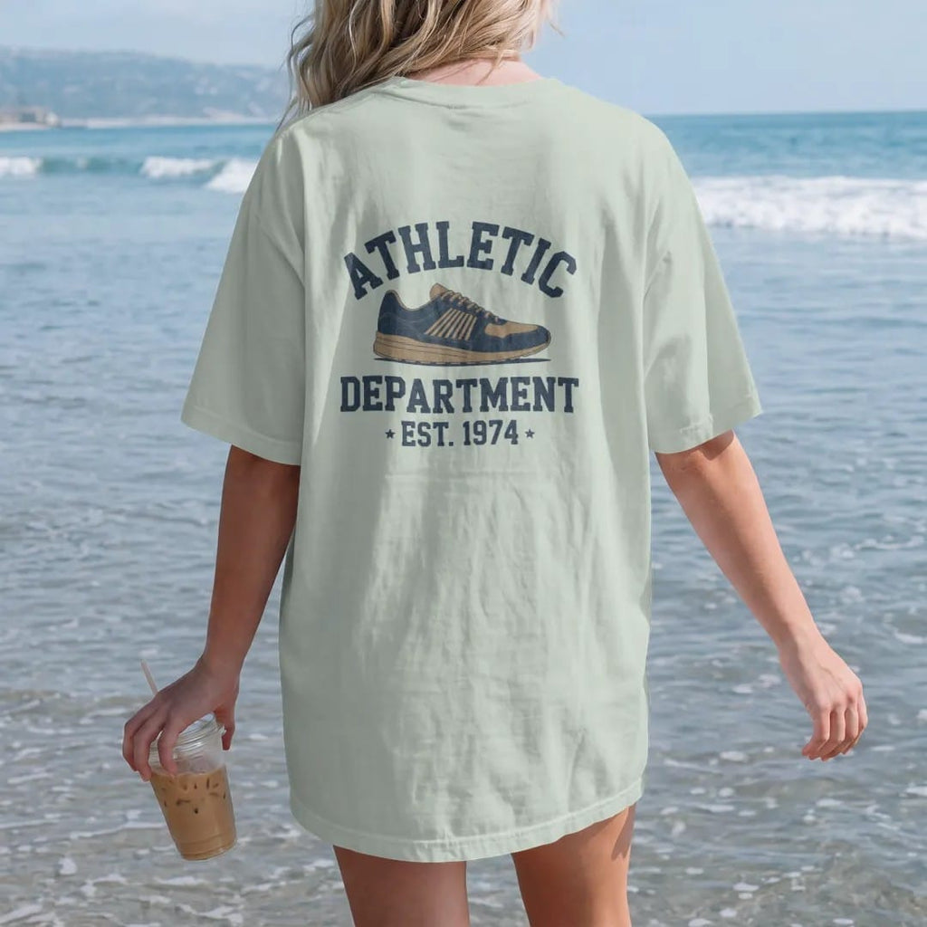 Woman on the beach in a Bay tee, back view: a vintage running shoe with speed lines, with "ATHLETIC" and "DEPARTMENT" and "EST. 1974" in chunky faded collegiate block letters, printed in faded varsity navy, washed red, mustard gold and pine green…