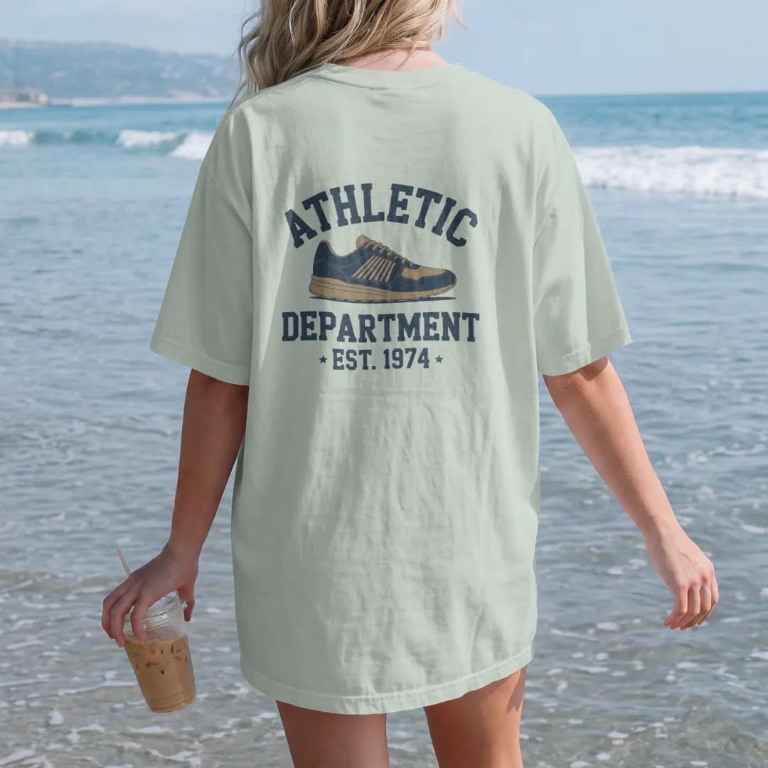 Woman on the beach in a Bay tee, back view: a vintage running shoe with speed lines, with "ATHLETIC" and "DEPARTMENT" and "EST. 1974" in chunky faded collegiate block letters, printed in faded varsity navy, washed red, mustard gold and pine green…