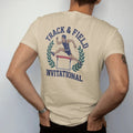 Man wearing a Sandstone tee, seen from the back: a runner clearing a hurdle mid-stride, framed by laurel leaves, with "TRACK & FIELD" and "INVITATIONAL" in chunky faded collegiate block letters, printed in faded varsity navy, washed red, mustard…