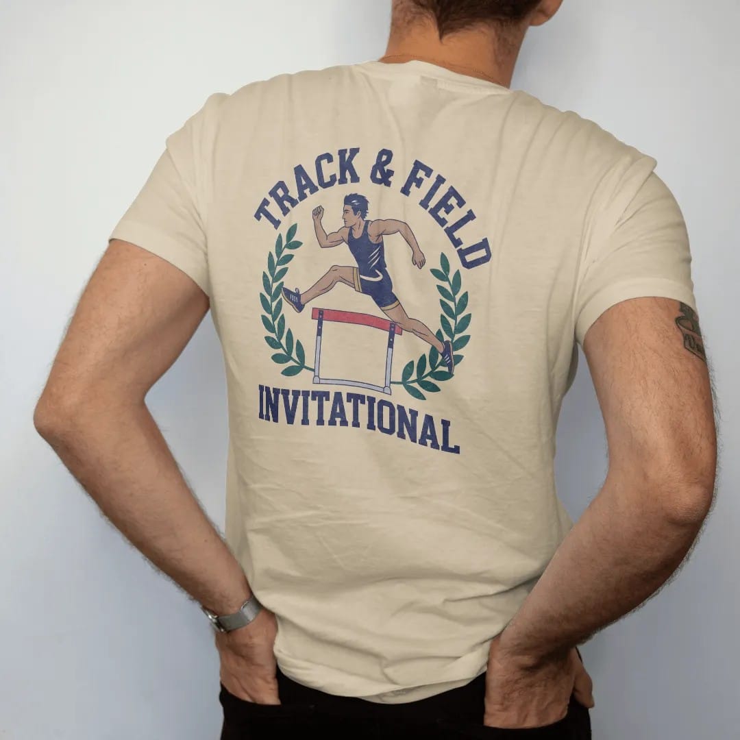 Man wearing a Sandstone tee, seen from the back: a runner clearing a hurdle mid-stride, framed by laurel leaves, with "TRACK & FIELD" and "INVITATIONAL" in chunky faded collegiate block letters, printed in faded varsity navy, washed red, mustard…