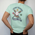 Man wearing a Chalky Mint tee, seen from the back: a stitched baseball over crossed wooden bats and a leather glove, with "SPRING TRAINING" and "BASEBALL CAMP" in chunky faded collegiate block letters, printed in faded varsity navy, washed red,…