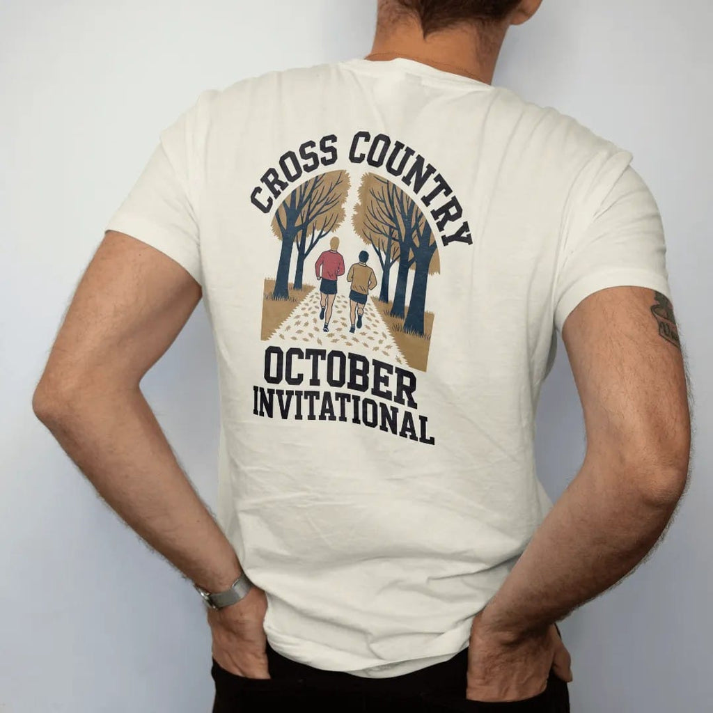 Man wearing a Ivory tee, seen from the back: two runners on a leaf-covered trail through autumn trees, with "CROSS COUNTRY" and "OCTOBER INVITATIONAL" in chunky faded collegiate block letters, printed in faded varsity navy, washed red, mustard gold…