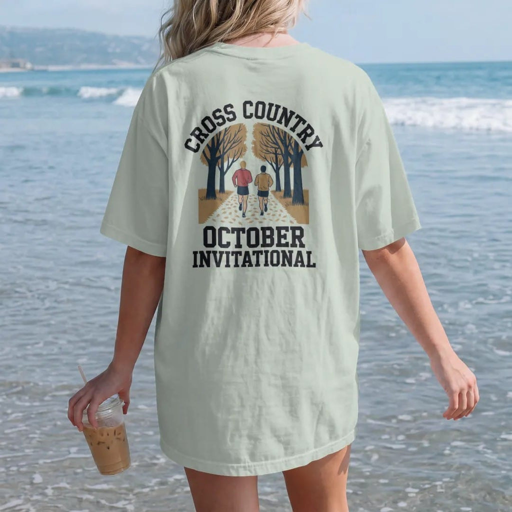 Woman on the beach in a Bay tee, back view: two runners on a leaf-covered trail through autumn trees, with "CROSS COUNTRY" and "OCTOBER INVITATIONAL" in chunky faded collegiate block letters, printed in faded varsity navy, washed red, mustard gold…