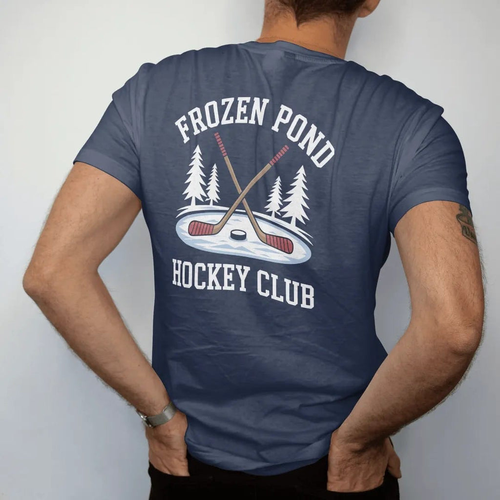 Man wearing a Navy tee, seen from the back: two crossed wooden hockey sticks and a puck on a frozen pond with snowy pines, with "FROZEN POND" and "HOCKEY CLUB" in chunky faded collegiate block letters, printed in faded bleacher cream, mustard gold,…