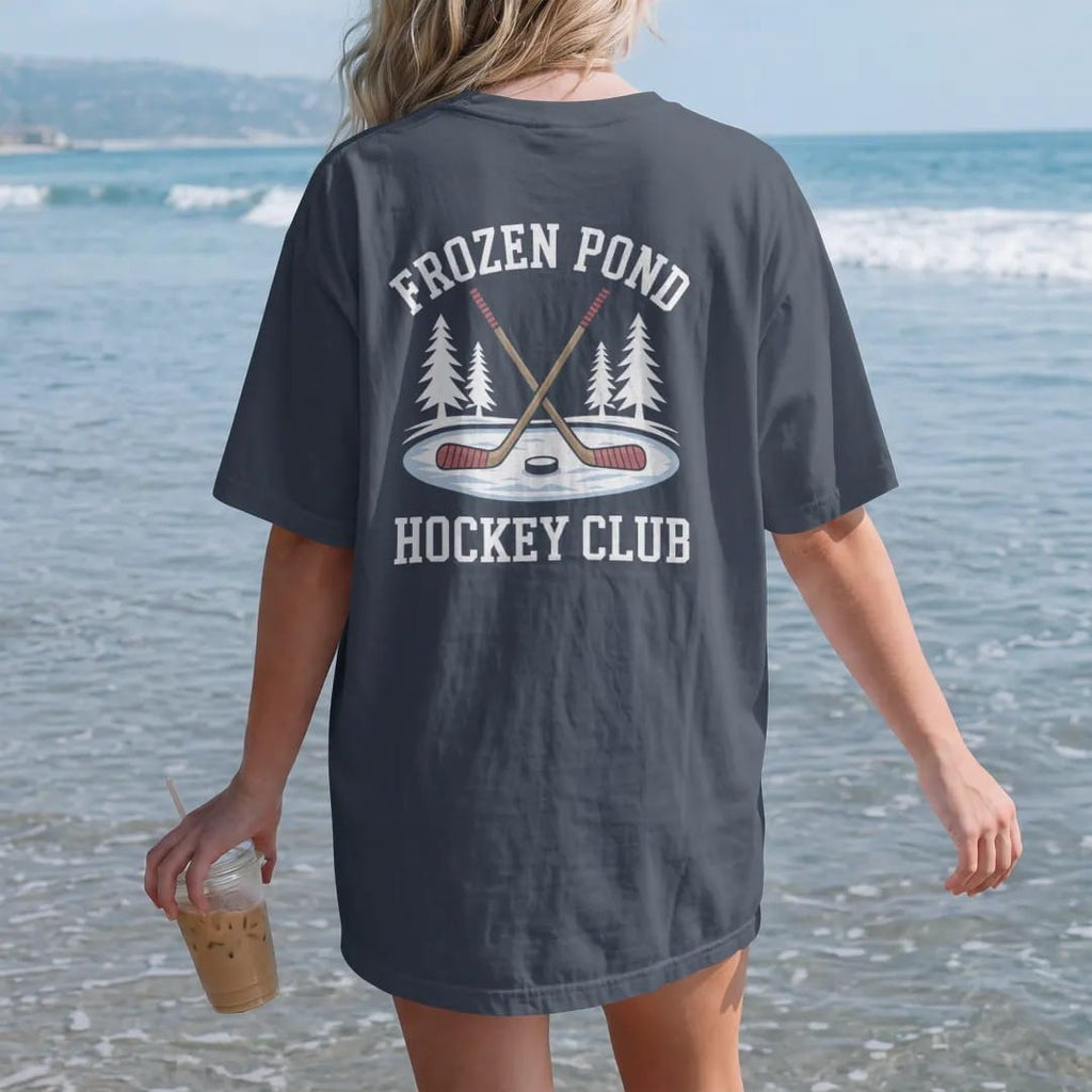 Woman on the beach in a Midnight tee, back view: two crossed wooden hockey sticks and a puck on a frozen pond with snowy pines, with "FROZEN POND" and "HOCKEY CLUB" in chunky faded collegiate block letters, printed in faded bleacher cream, mustard…