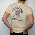 Man wearing a Sandstone tee, seen from the back: a wooden tennis racquet and a fuzzy ball with motion lines, with "MATCH POINT" and "TENNIS CLUB" in chunky 1970s swash script, printed in faded varsity navy, washed red, mustard gold and pine green…