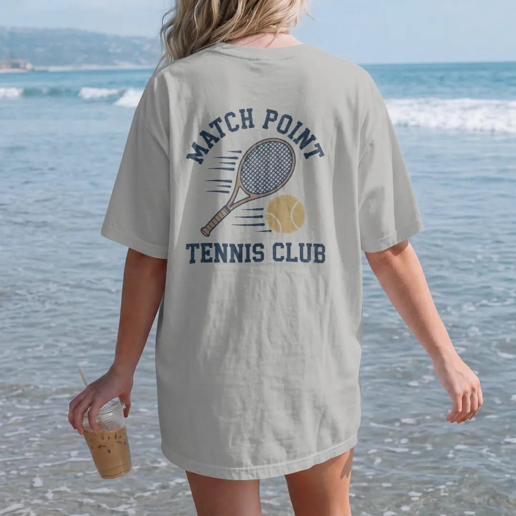 Woman on the beach in a Granite tee, back view: a wooden tennis racquet and a fuzzy ball with motion lines, with "MATCH POINT" and "TENNIS CLUB" in chunky 1970s swash script, printed in faded varsity navy, washed red, mustard gold and pine green…