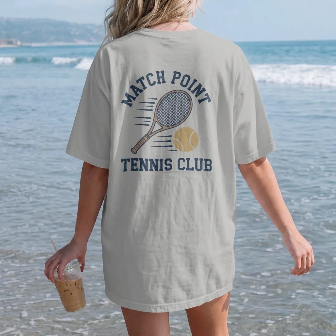 Woman on the beach in a Granite tee, back view: a wooden tennis racquet and a fuzzy ball with motion lines, with "MATCH POINT" and "TENNIS CLUB" in chunky 1970s swash script, printed in faded varsity navy, washed red, mustard gold and pine green…